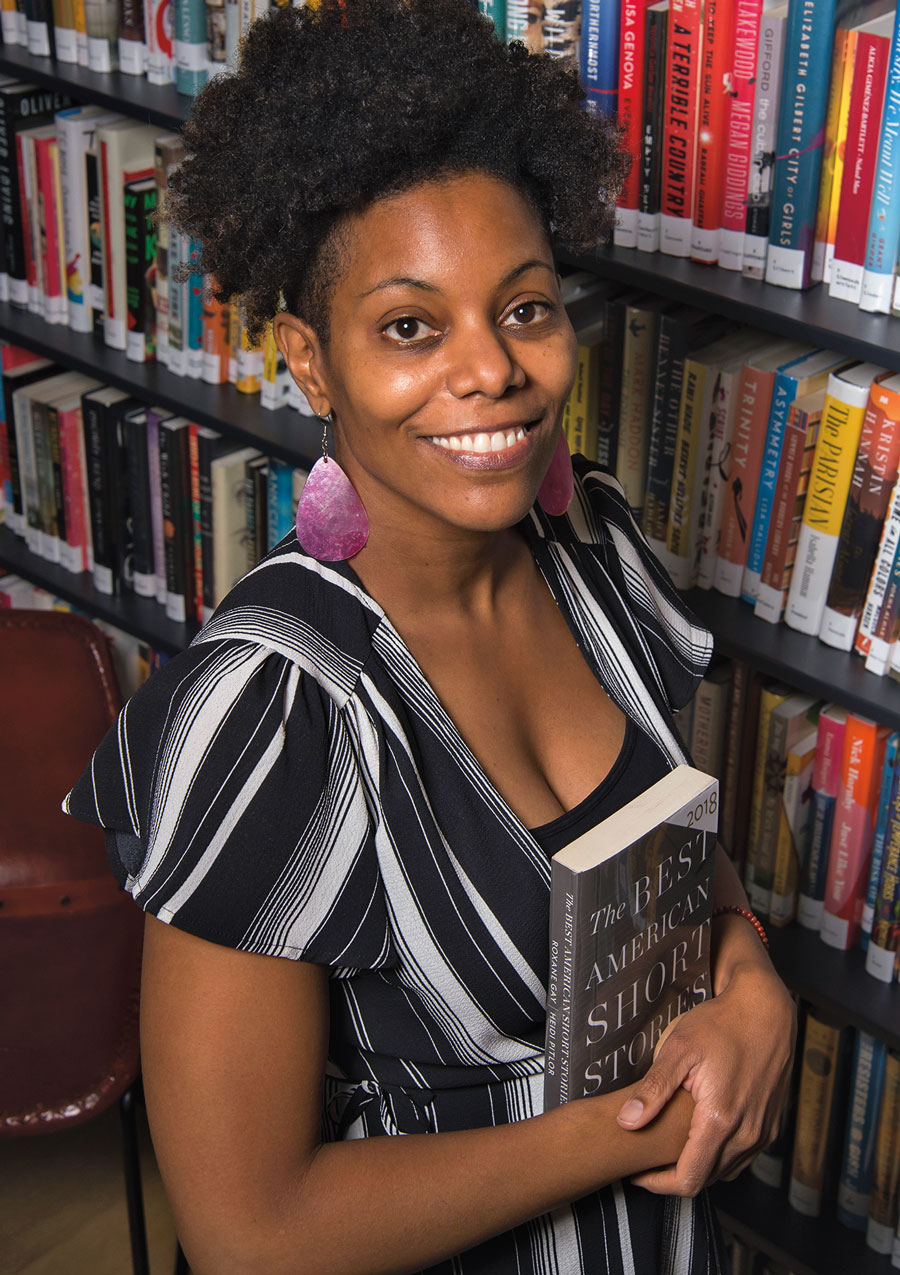 Kim Foote holding a book in a library, smiling and wearing a black-and-white top and purple earrings