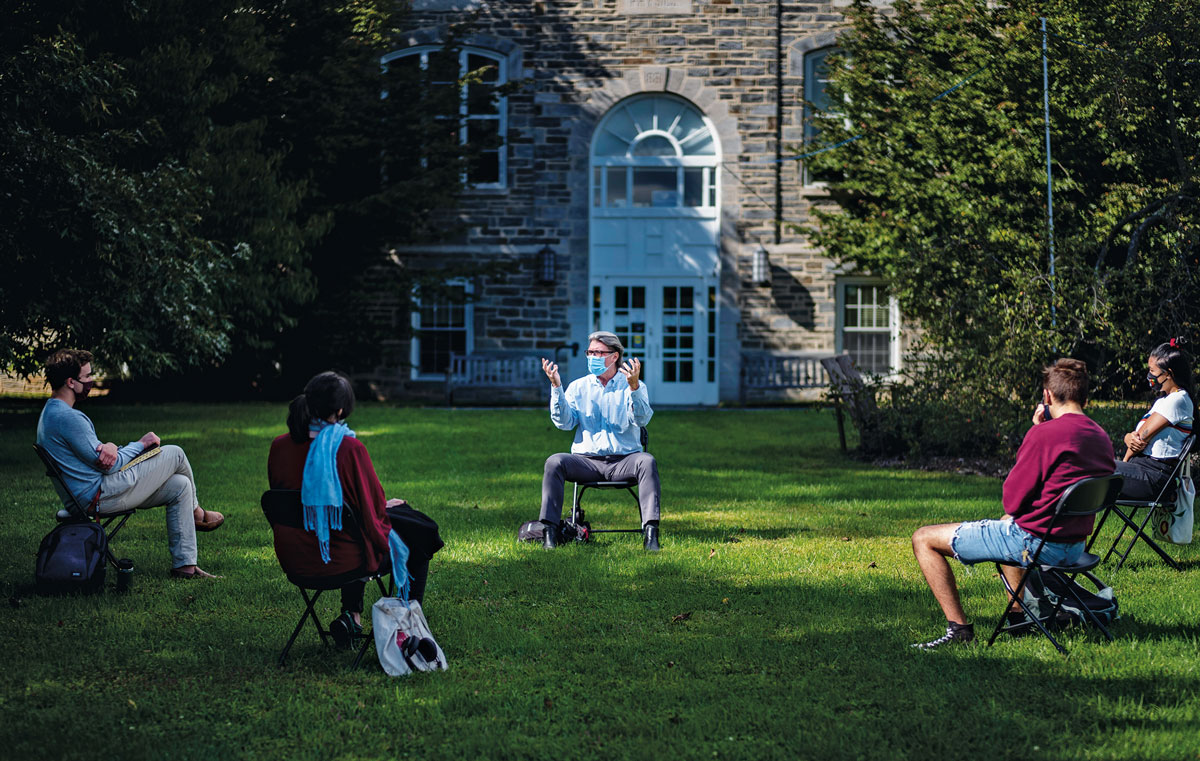 Mark Wallace, sitting outside in a courtyard on Swarthmore’s campus, wearing a mask while speaking and gesturing to four students, masked and physically distanced in a semicircle