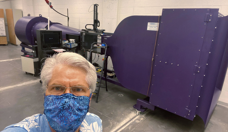 Carr Everbach, wearing glasses and a blue patterned mask, with a large purple machine in the background