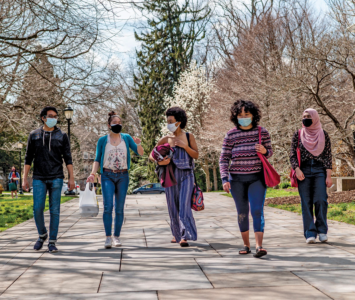 1 male and four female students walking in line through campus on a sunny day, with budding trees in the background. They are all wearing facemasks.