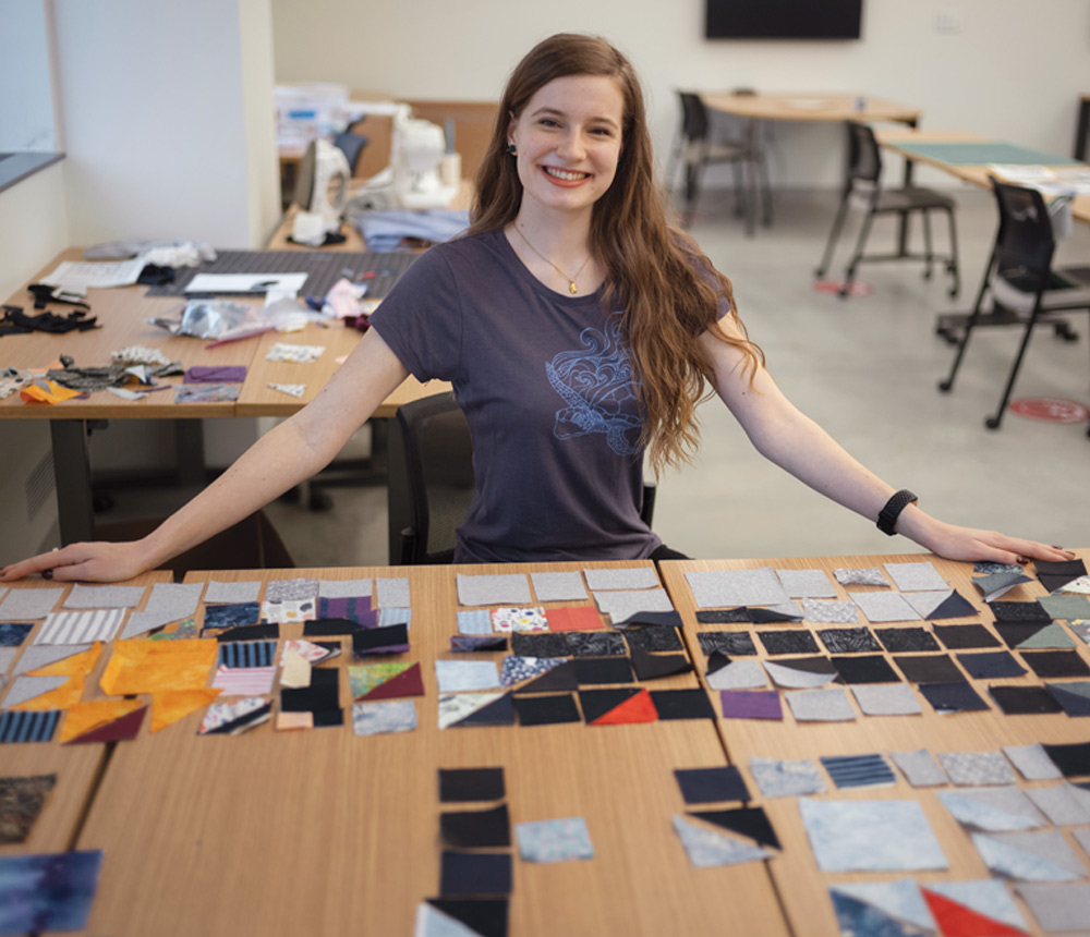 Hannah Watkins sitting inside at a table that is lined with squares of fabric for her quilt. She is smiling and wearing a dark-gray T-shirt.