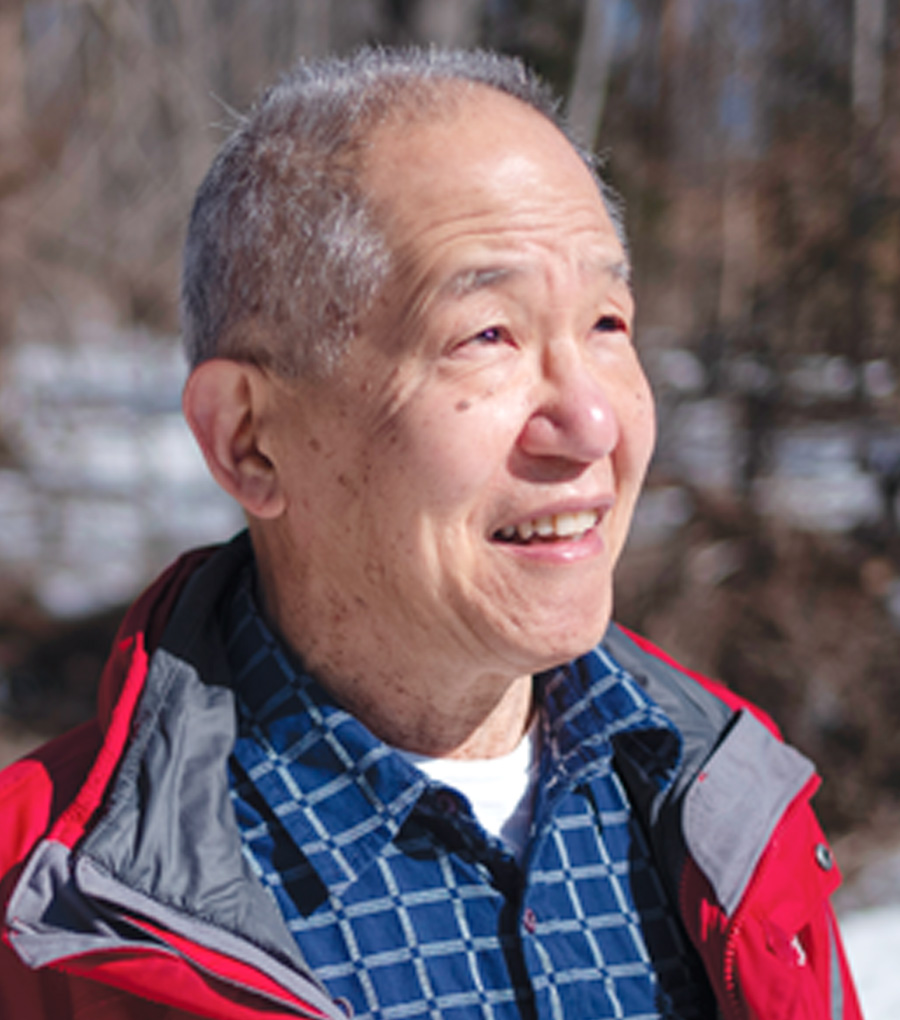 Headshot of Leonard Nakamura, photographed outside with snow on the ground. He’s wearing a navy button-down and a red coat, and the sun is shining on his face.