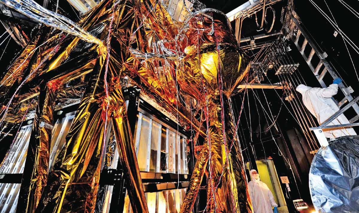 A golden metallic structure with wires hanging from it. Two people dressed in white protective gear stand next to it.