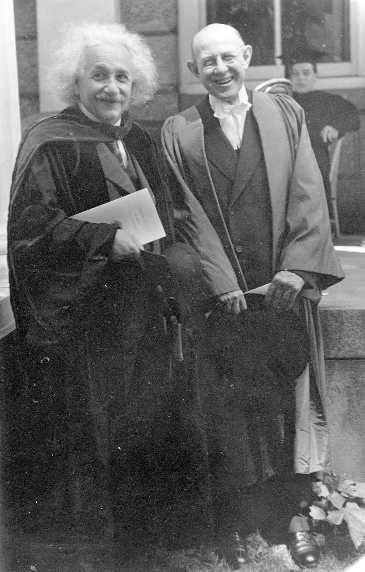 Albert Einstein and Frank Aydelotte, standing together wearing commencement regalia. Einstein is holding a commencement program. In the background is a student (Eugene Lang) sitting in a chair.
