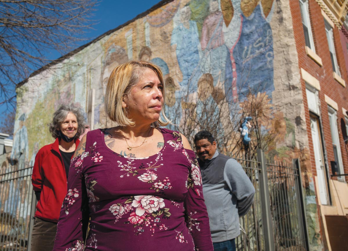 Peaches Ramos Bautista wearing a purple blouse with flowers, standing next to a rowhouse with a mural painted on its side. Behind her are Jean Murdock Warrington, wearing a red jacket, and Hector Colon, dressed in a gray jacket.