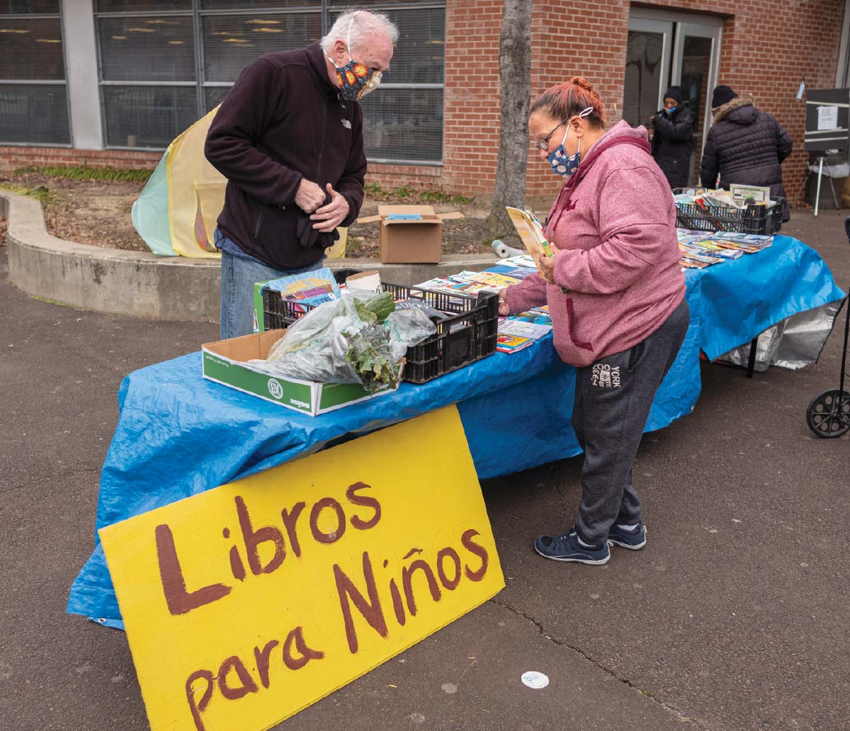 Peter Warrington, wearing a black jacket and a facemask, looking down at a table of books and vegetables. A woman dressed in a pink jacket, gray sweatpants, and a facemask, stands on the other side of the table holding a few books. A yellow sign next to the table reads “Libros para Niños.”