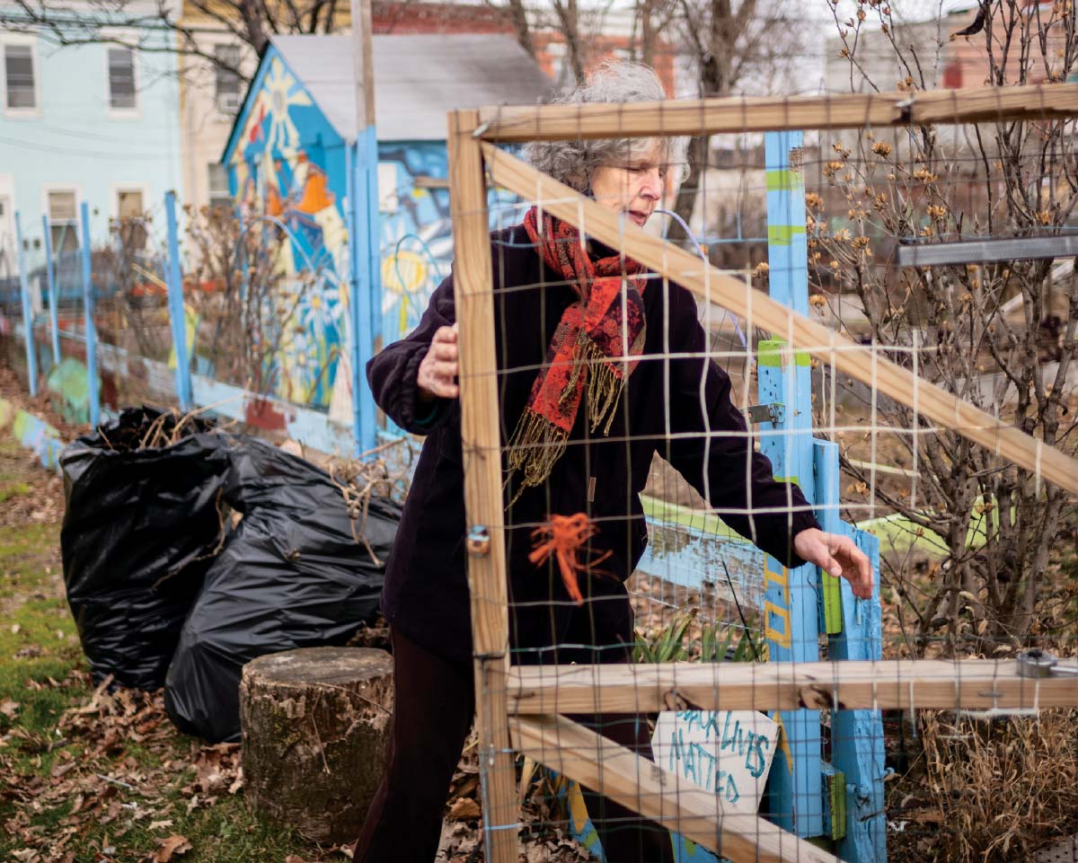 Jean Murdock Warrington, wearing a black jacket and red scarf, opens a wooden gate to the garden. A light-blue fence and colorfully painted shed are in the background.