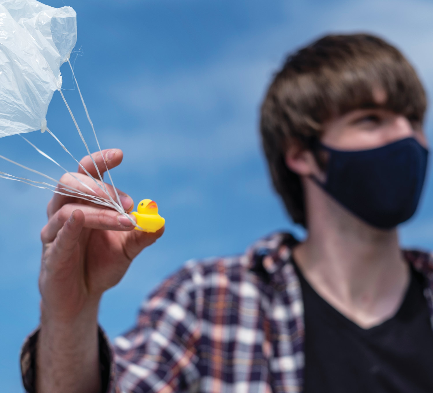 Zane Meyer wearing a black V-neck T-shirt, a flannel button-down, and a black facemask, holding a small, yellow rubber duck that has a plastic parachute attached to it. Behind them is a bright blue sky.