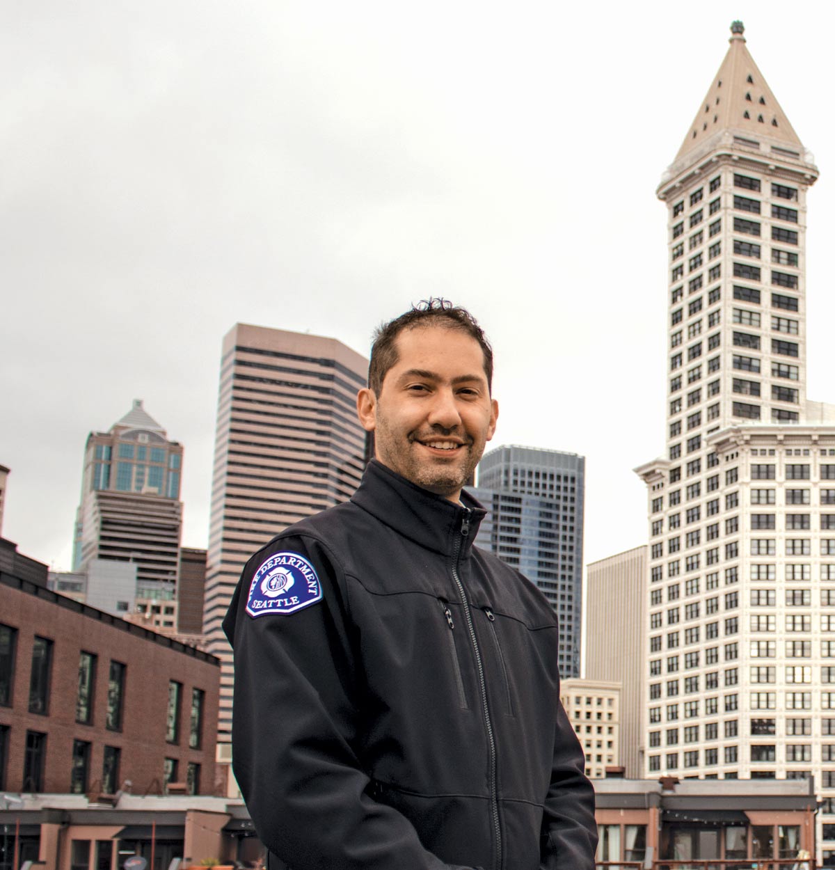 Jon Ehrenfeld outside in a downtown area with buildings in the background. He’s wearing black jacket with a “Fire Department Seattle” patch on the arm.