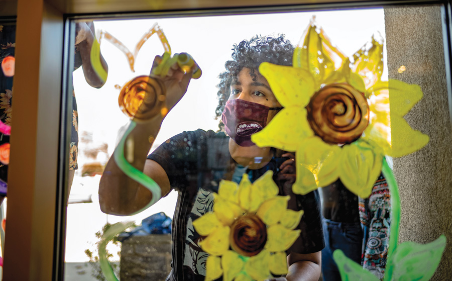 Shot from the other side of the window, Amy Marcalle paints images of sunflowers while wearing a Swarthmore facemask and a black T-shirt. 
