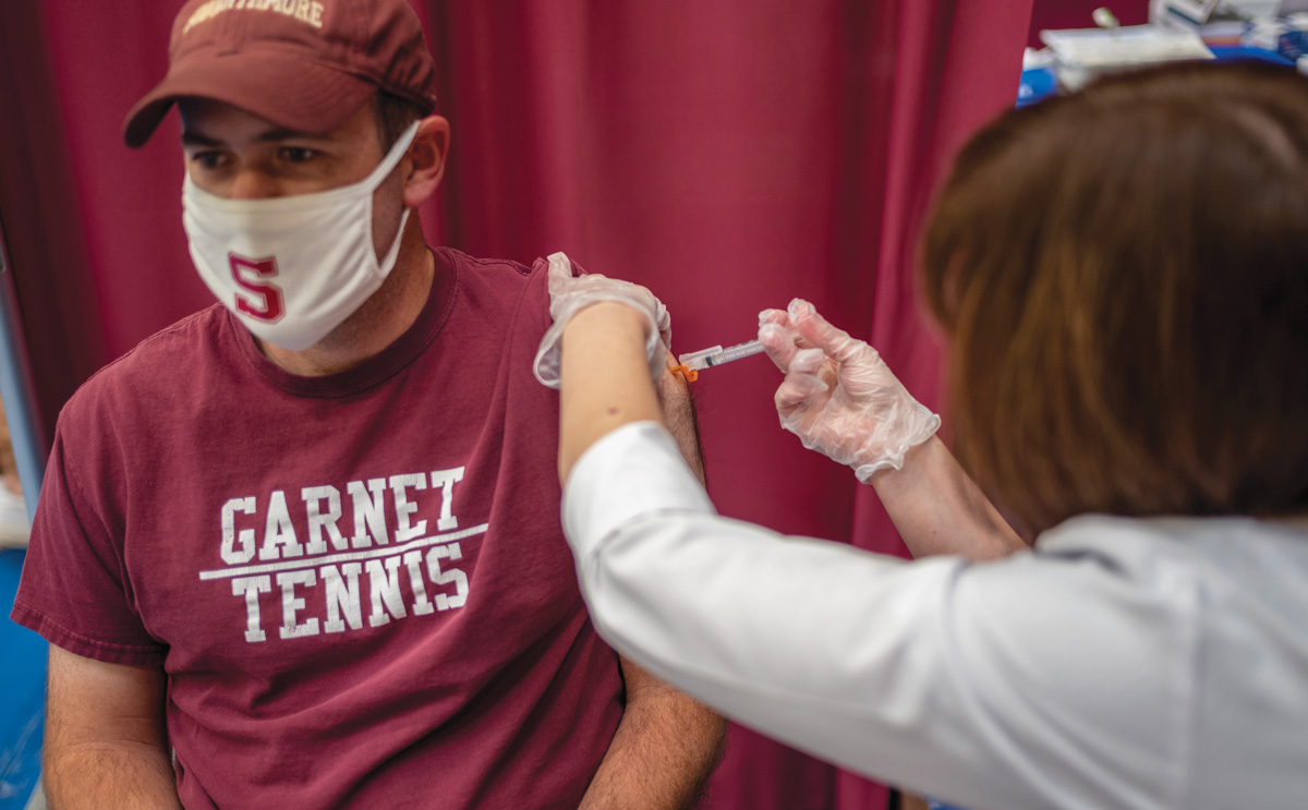 Jason Box receiving a vaccine from a female health care worker. Jason is wearing a Garnet Tennis T-shirt, a garnet-colored Swarthmore hat, and a white facemask with the Swarthmore “S” on it. The woman has on a white lab jacket and medical gloves.