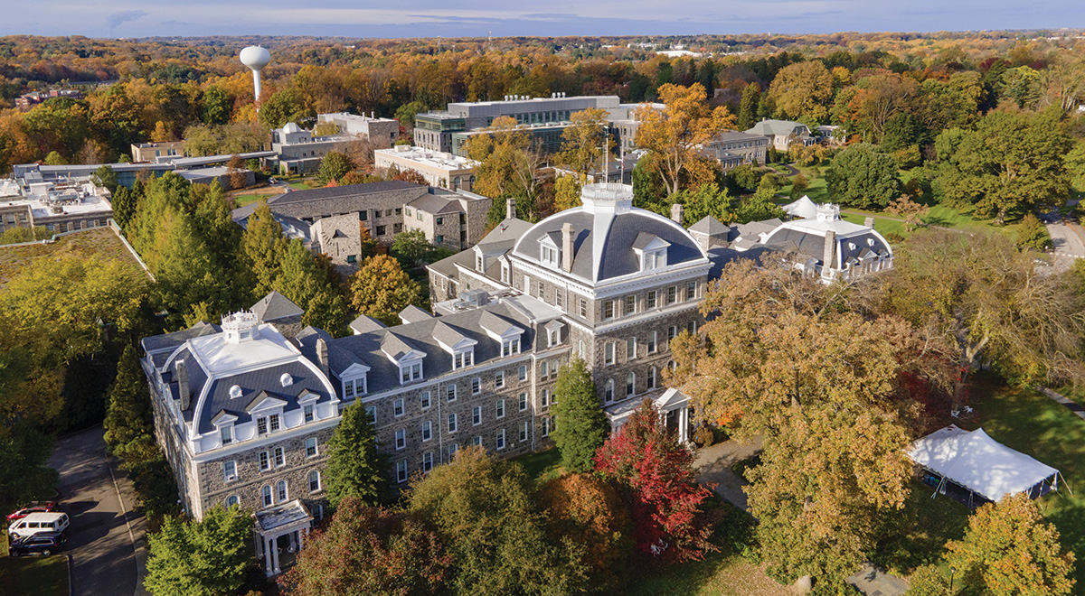 A drone shot of Swarthmore’s campus on a sunny day. Parrish Hall is in the center, surrounded by trees with green, yellow, red, and brown leaves.