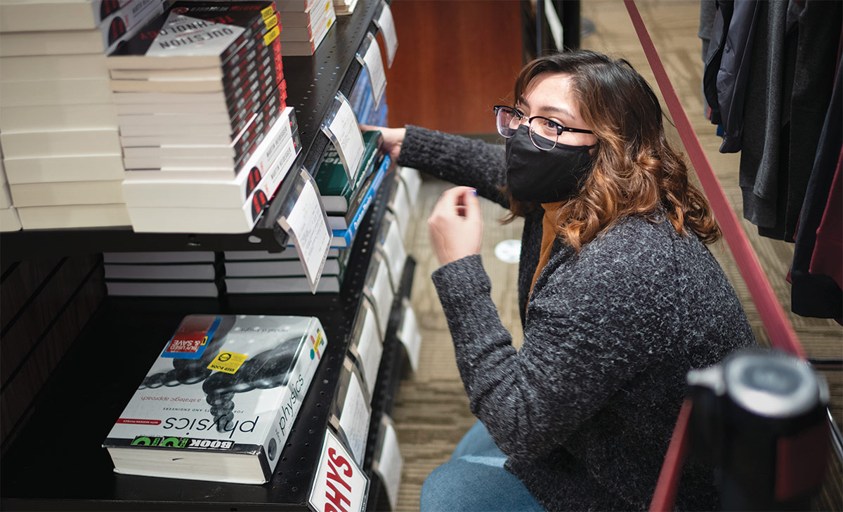 Edna Olvera, wearing glasses, a black facemask, and a gray sweater, squatting down near the floor while looking at stacks of textbooks on shelves in the bookstore.