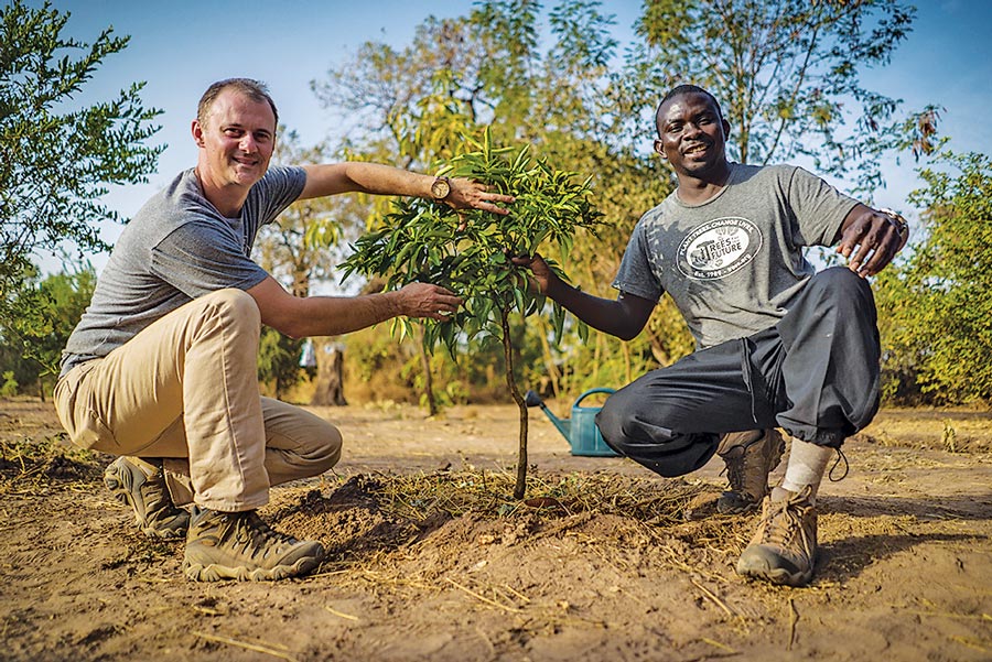John Leery and Omar Ndao kneeling on either side of a young, newly planted tree. Both men are wearing t-shirts, pants, and hiking boots.