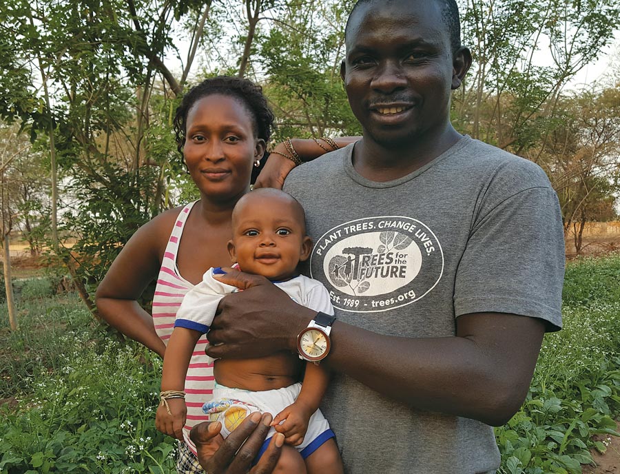 Omar Ndao, wearing a gray Trees for the Future shirt, holding his infant son, Cheikh, who is smiling. Wife Fama has her arm on Omar’s shoulder and is wearing a red-and-white striped tank top.