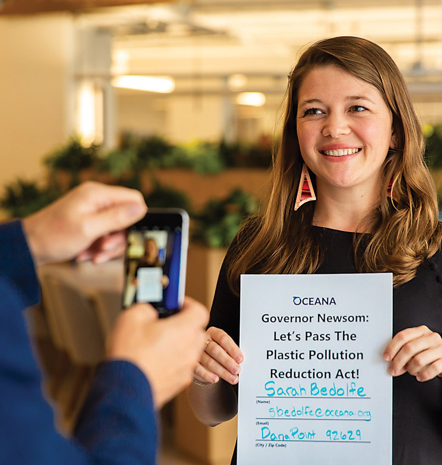 Sarah Bedolfe smiling as someone takes her photo with a phone. She’s holding a sheet of paper that reads, “Oceana. Governor Newsom: Let’s Pass The Plastic Pollution Reduction Act!”