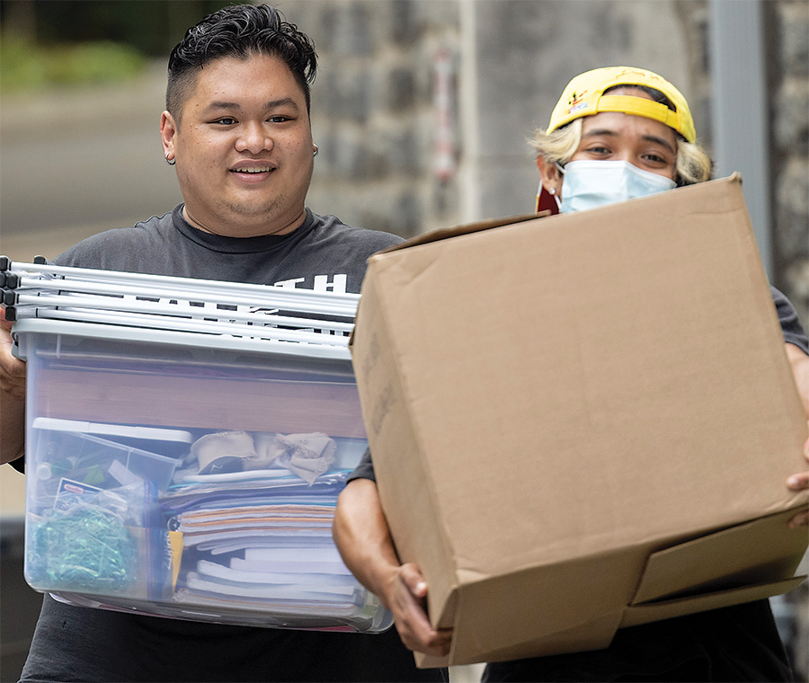 Two students on move-in day, one carrying a large plastic bin, the other a large brown box. One student is smiling, and the other is wearing a facemask and a backward yellow ballcap