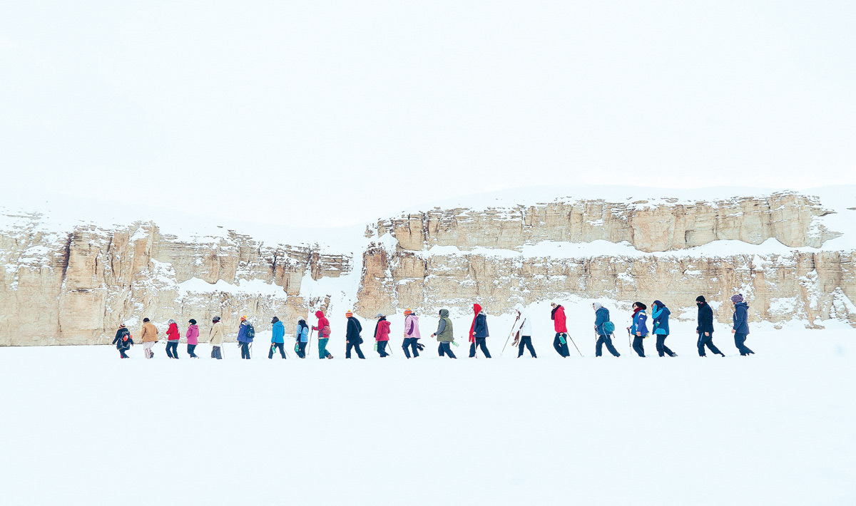 A line of people trekking through snow, with cliffs in the background.