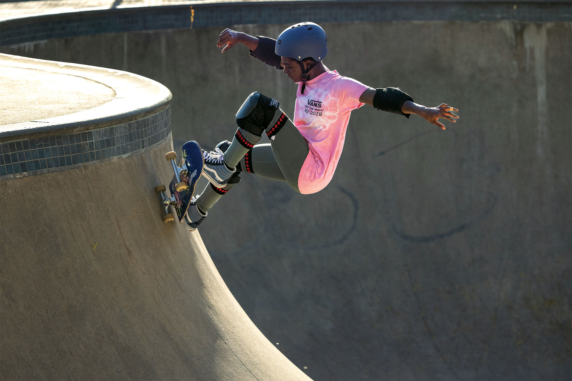 Mame Bonsu, wearing a pink tie-dye Vans shirt and protective gear, skateboarding in a skatepark.