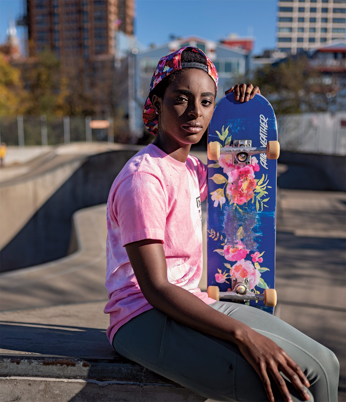 Mame Bonsu, wering a pink tie-dye t-shirt and multicolored ballcap, turned backward, sits on a ledge in a skatepark, holding her blue floral skateboard