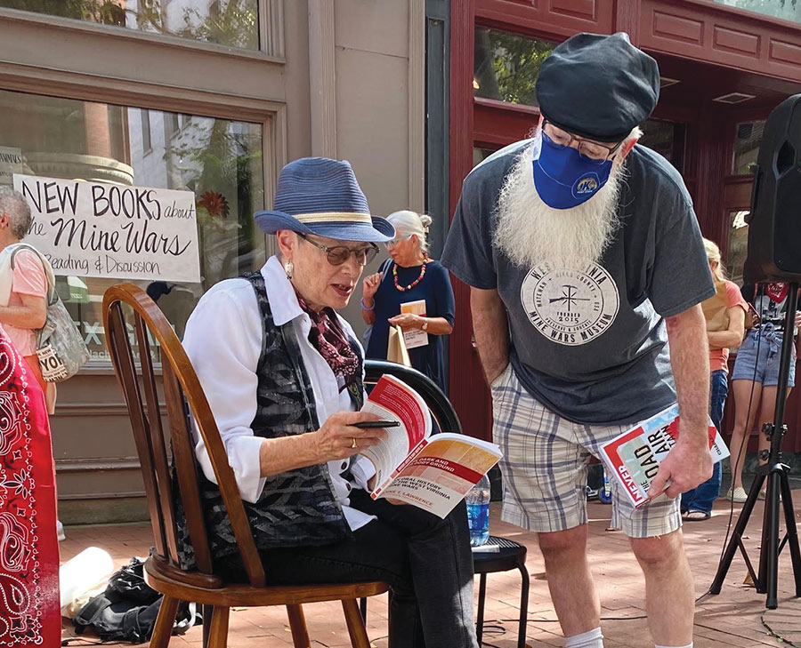 Anne T. Lawrence, sitting in a brown chair outdoors, wearing a blue fedora and holding a book. She is speaking with a man in a blue mask with a long, white beard, wearing a green beret and t-shirt