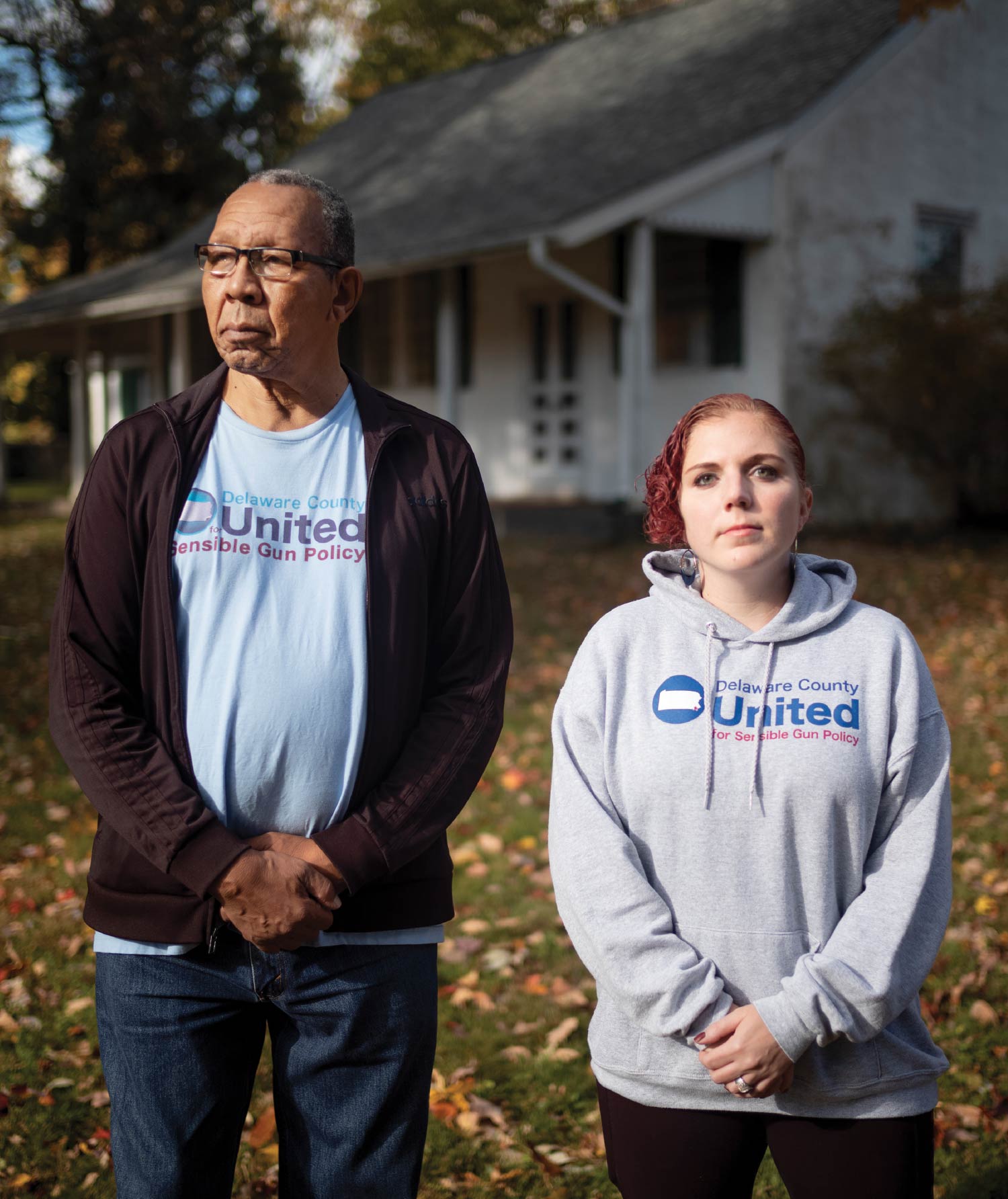 John Linder and Jess Frankl, wearing stoic looks and attire featuring the logo for Delaware County United for Sensible Gun Policy. They stand outside a Quaker meetinghouse with leave on the ground.