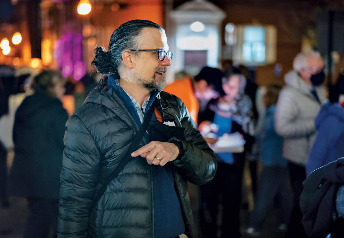 Lee Smithey, looking to the right while pointing left, sporting a ponytail, glasses, and a black puffer coat. He’s at an evening rally against gun violence