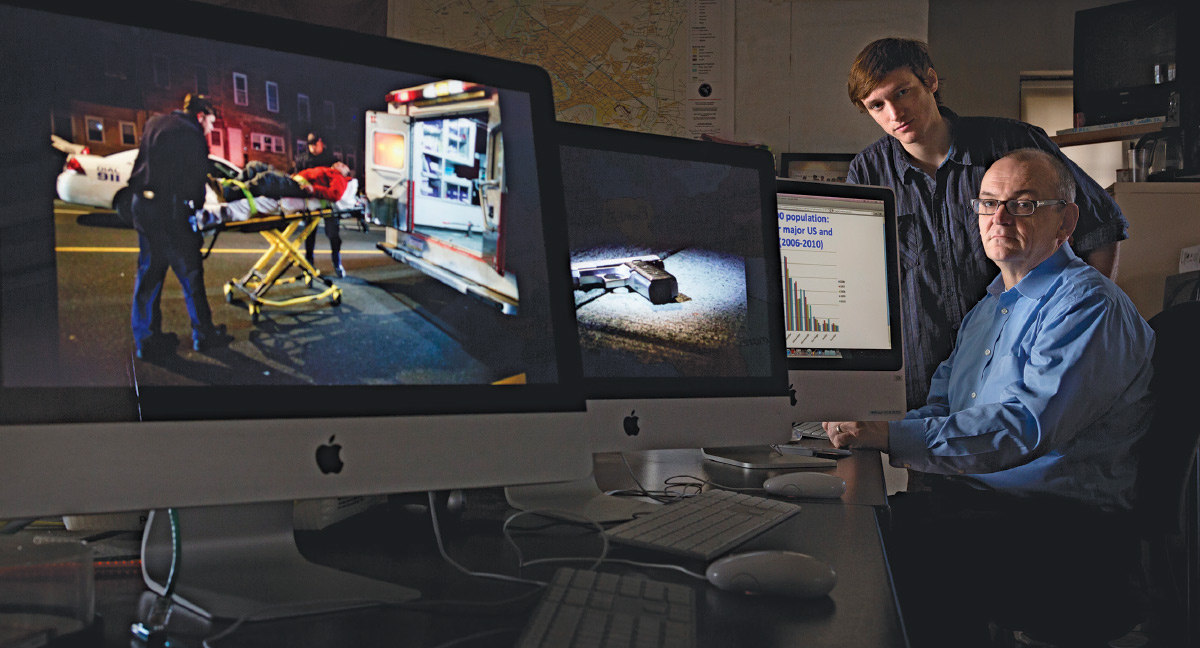 Jim MacMillan wearing a blue button-down, with student Aaron Moser behind him. Multiple computer monitors in the foreground display images related to gun violence.