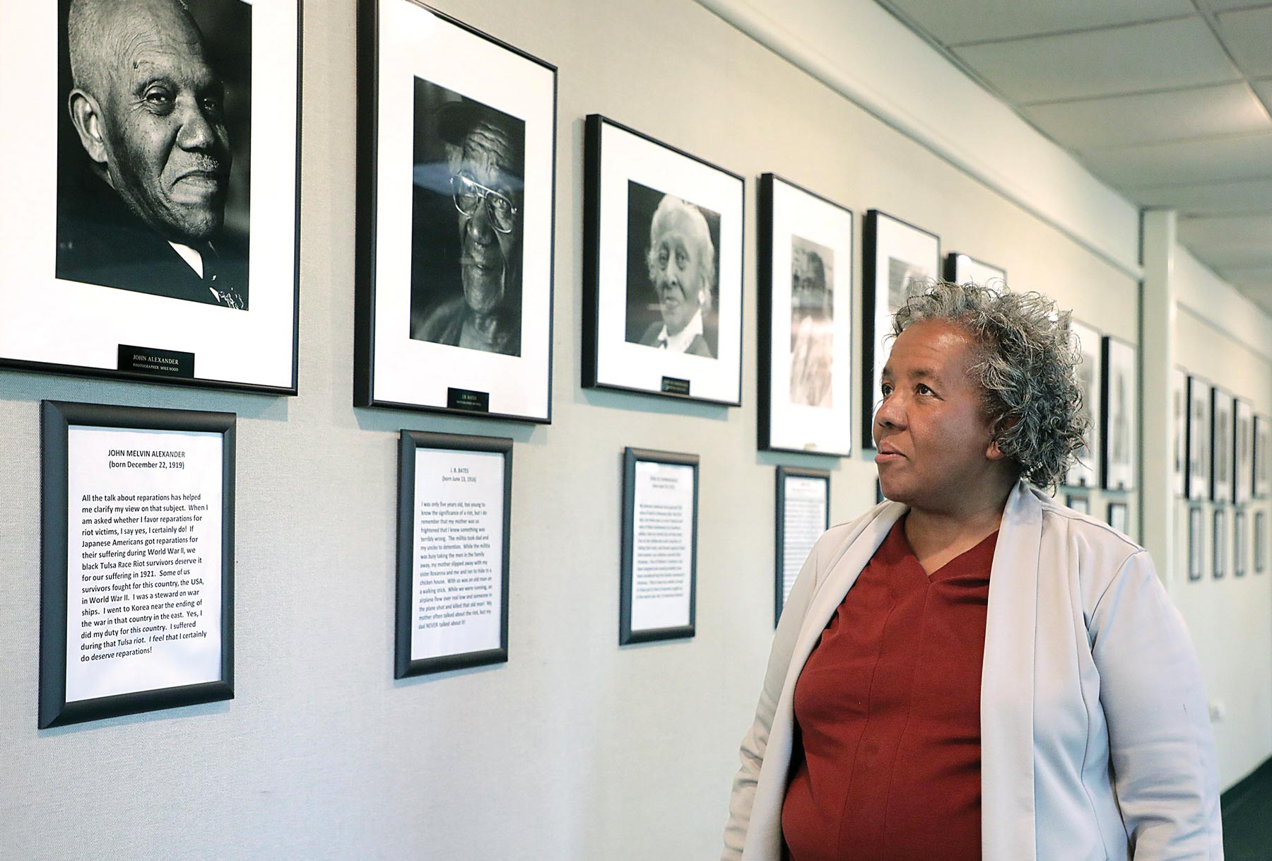 Sandra Alexander, wearing a maroon shirt and gray jacket, looking up at a black-and-white portrait of her father, John Melvin Alexander — one of many faces in a row of photographs