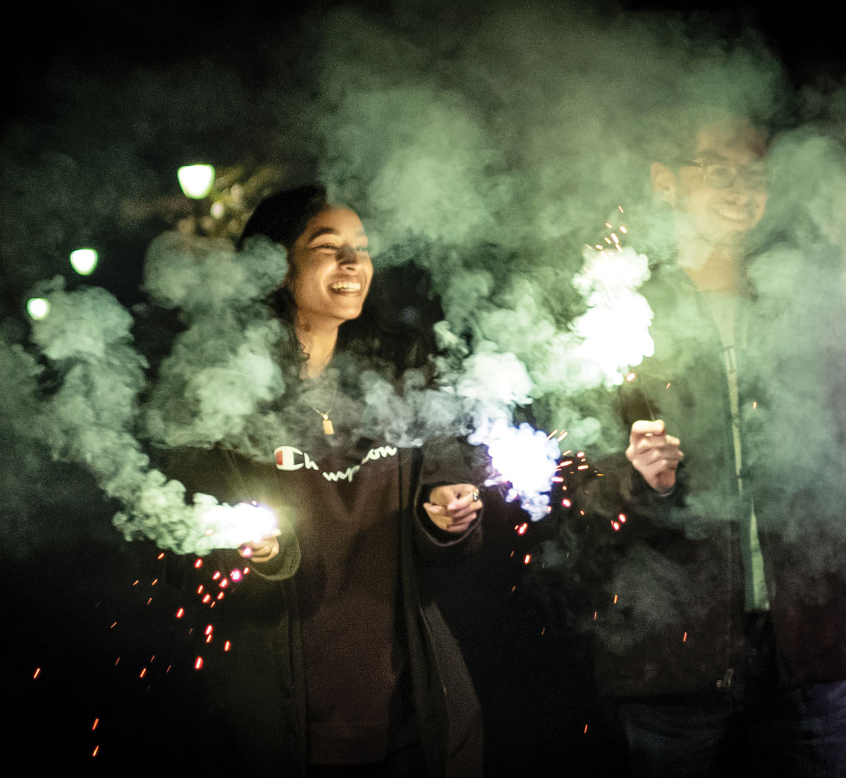 Two smiling students are surrounded by smoke as they hold sparklers outside at nighttime
