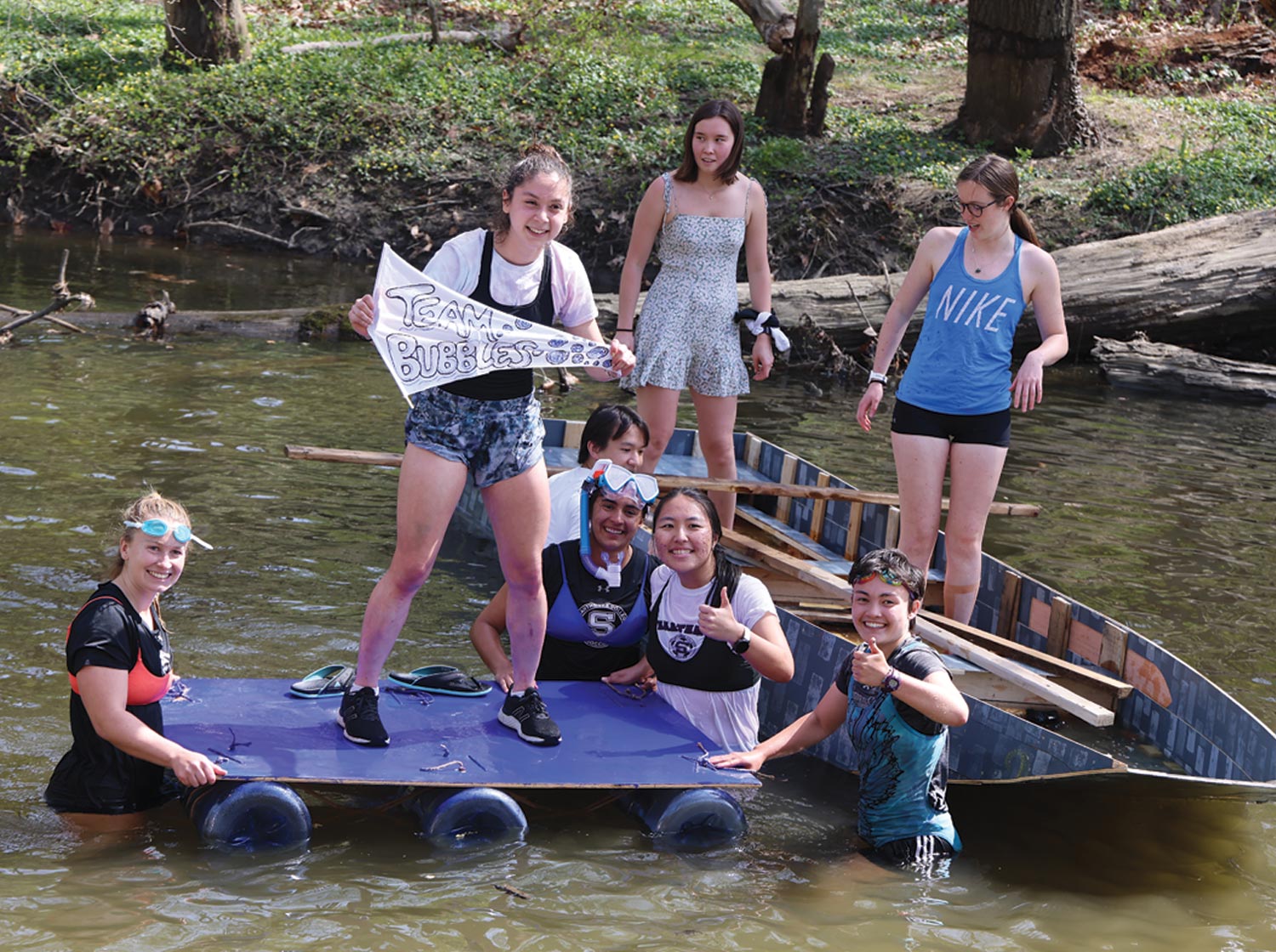 Young people on a raft in a creek hold flags and smile.