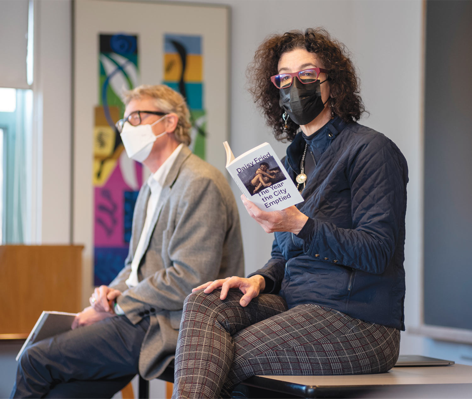 Two adults sit on a desk wearing masks. The woman with glasses reads from a book.