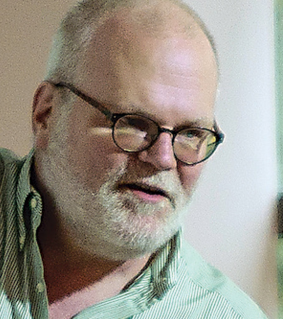 A bald man with white beard and glasses wears a green shirt.