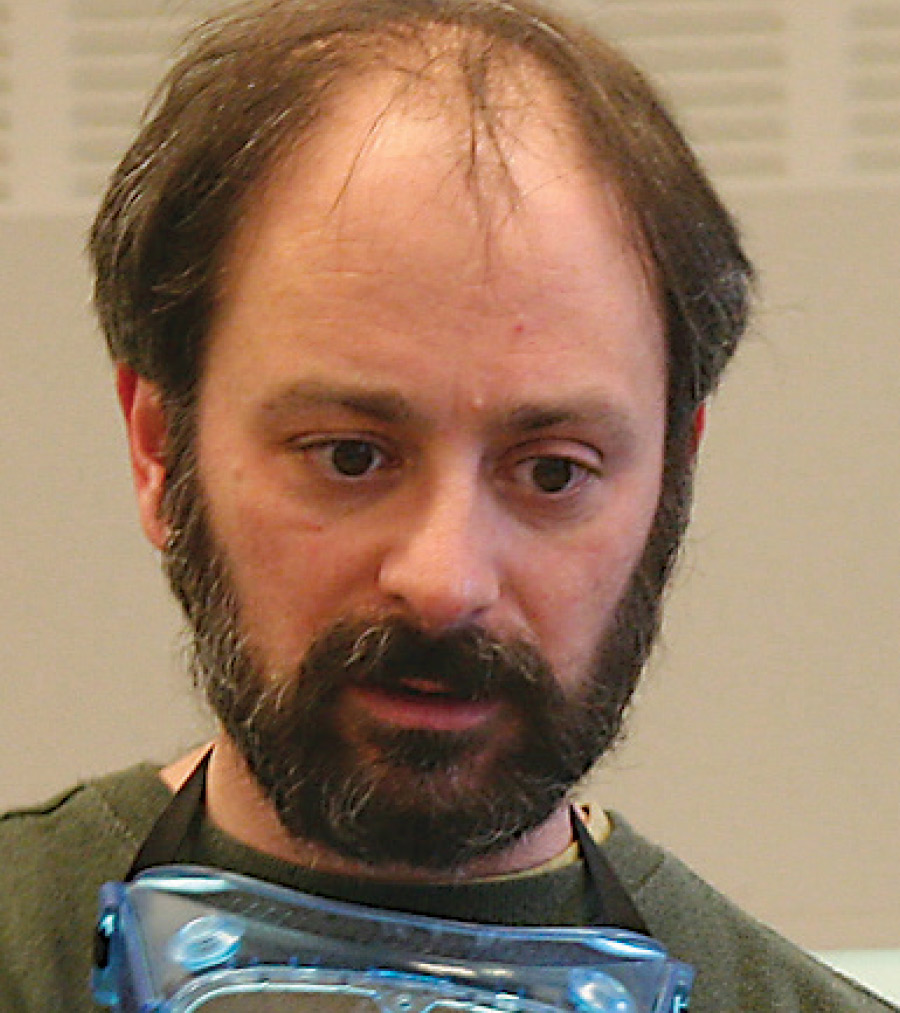 A concerned looking man with beard has goggles on his neck.