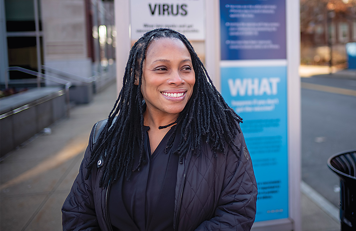 A woman with braids smiles in front of a bus stop with a blue sign.