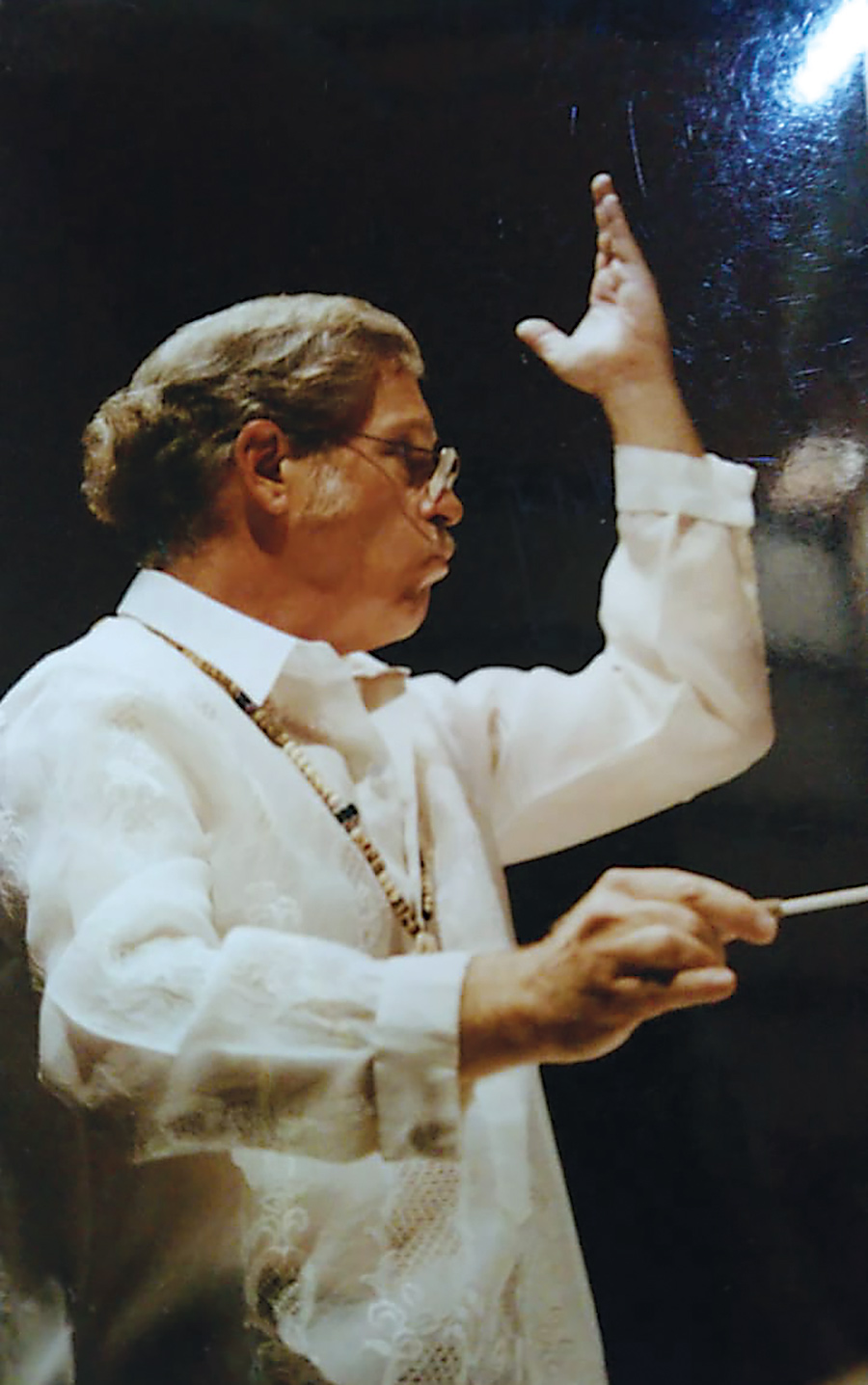 A man dressed in white dramatically holds his left arm in the air as he conducts an orchestra.