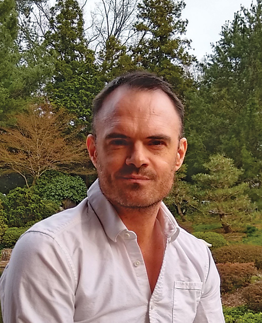 A man wearing a white shirt is in a garden setting with a serious expression.