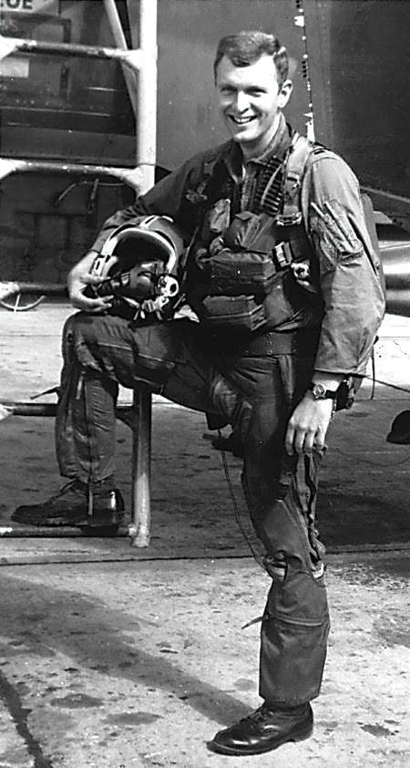 A man in a Air Force uniform smiles in front of a ladder leading to a jet. He holds a helmet.