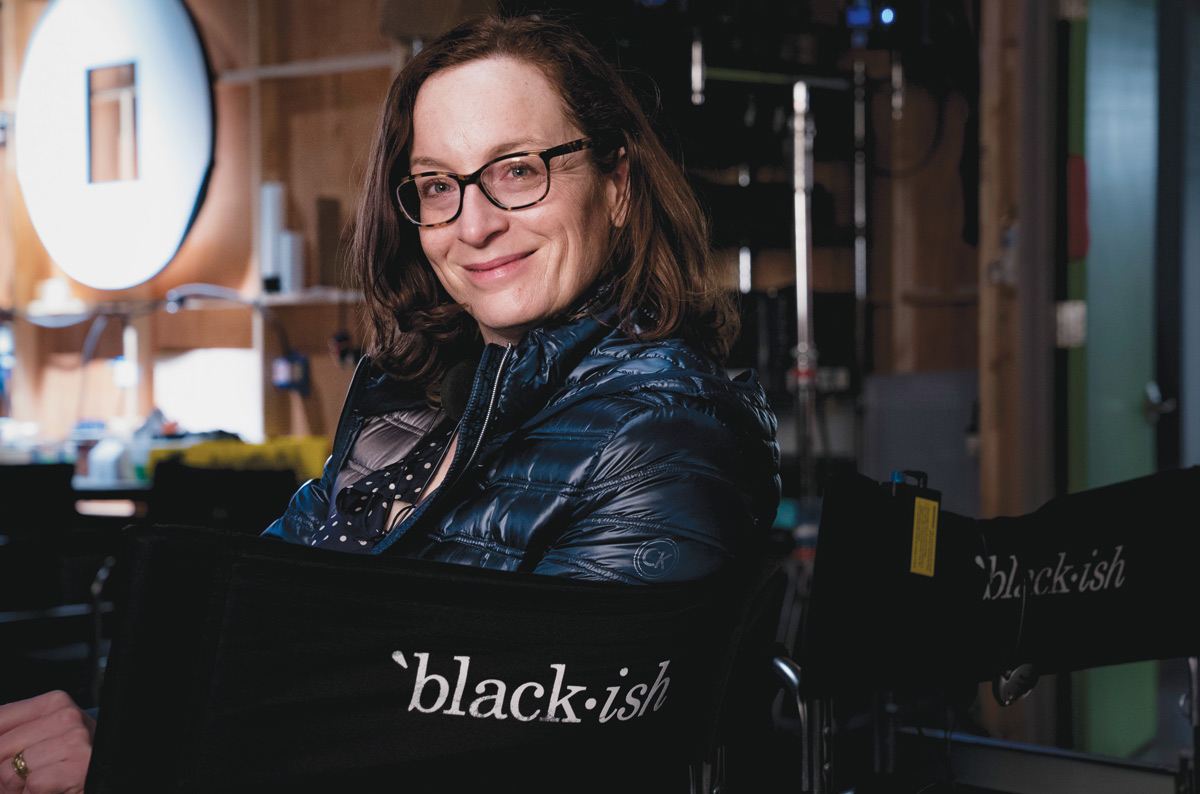 A woman with glasses and a puffy coat sits sideways in a chair that reads Blackish