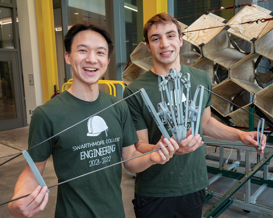 Two students wearing green "Swarthmore College Engineering" t-shirts smile at the camera while holding their invention. It's a wire frame shaped like the wings of a plane.