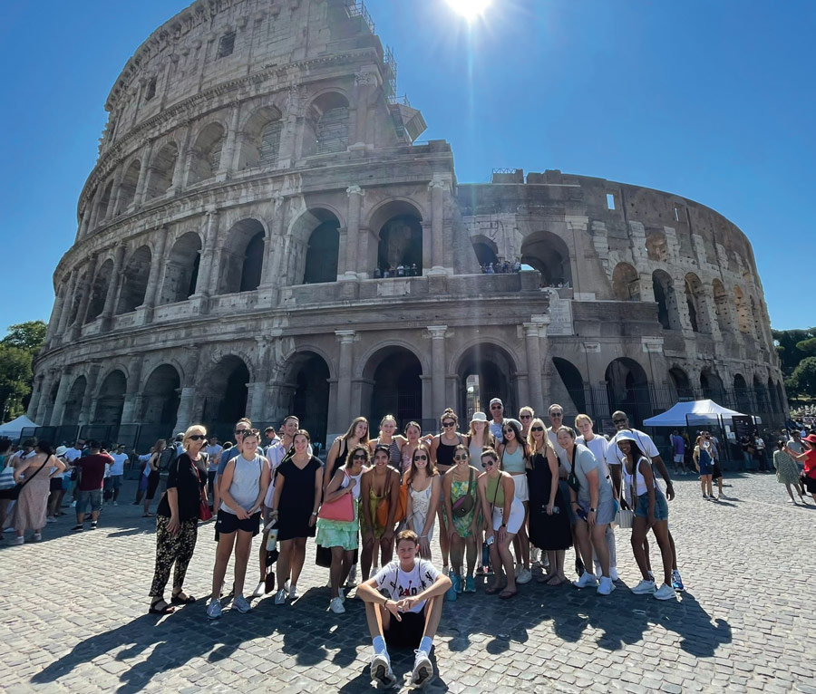 A group of students stand in front of the Colosseum in Rome on a sunny day.