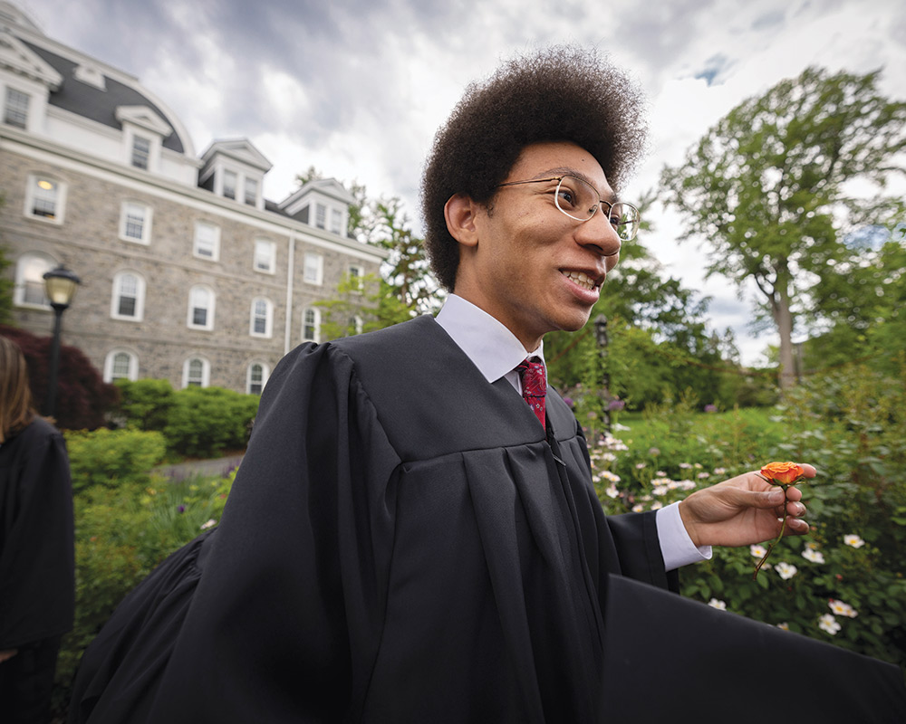 A student, wearing a black graduation robe and holding an orange flower, smiles.