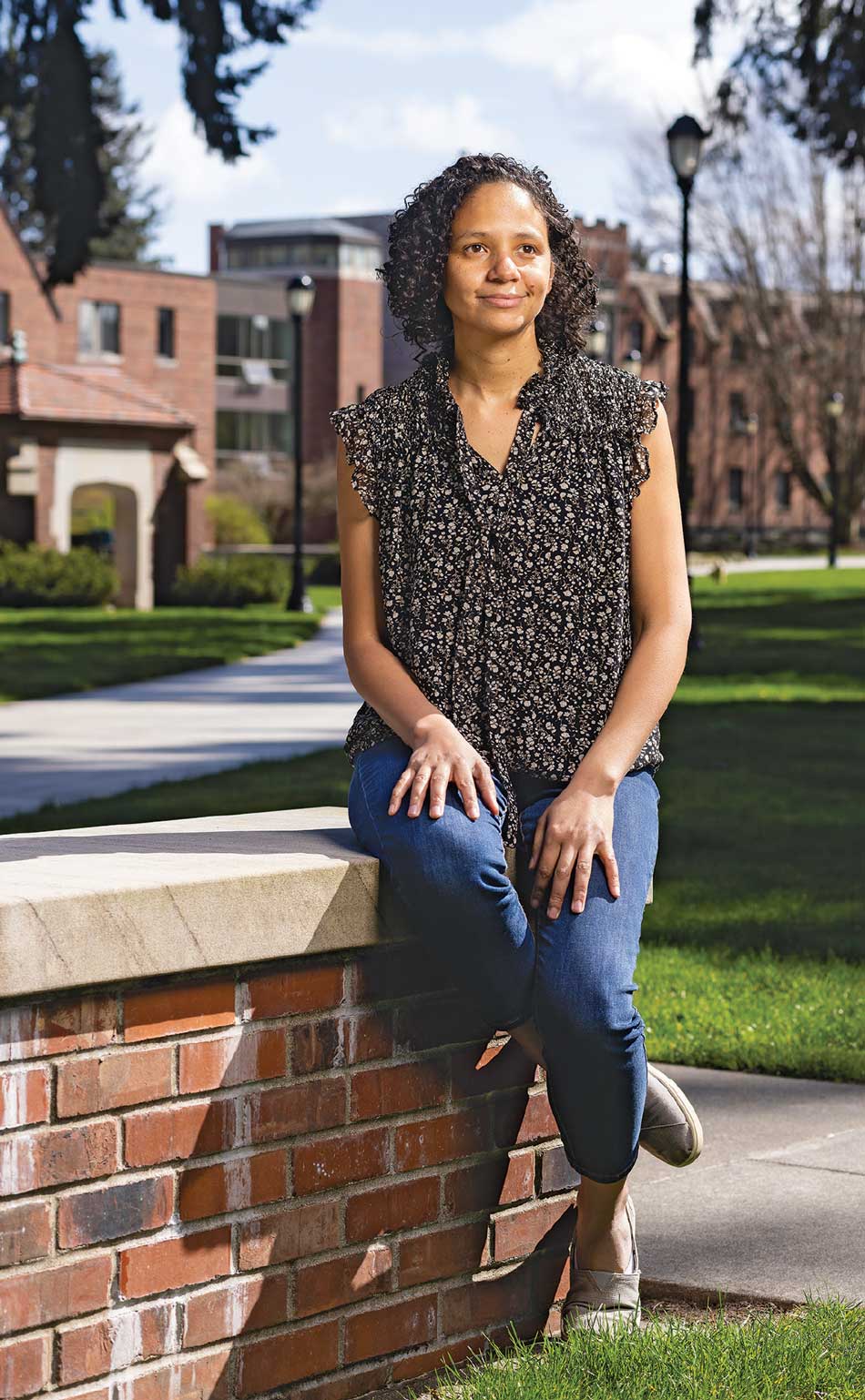 America Chambers poses sitting on a brick half-wall, wearing a black and white floral shirt and jeans. She appears to be on a college campus.