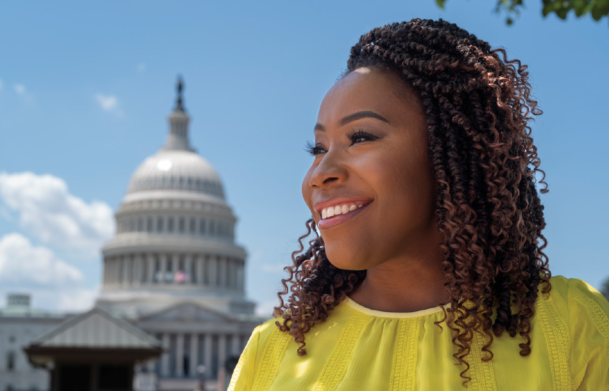 A Black woman reporter wearing a yellow shirt smiles in the foreground. The U.S. Capitol building and a blue sky are in the background.