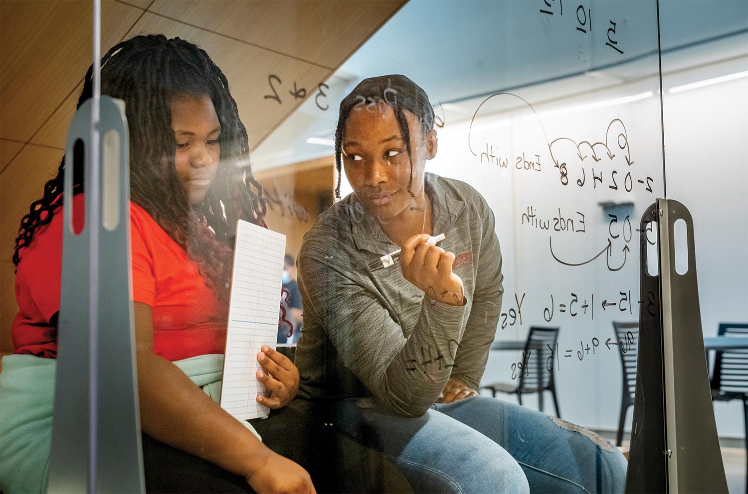 A college-age Black woman student teaches math to a younger Black woman student by writing equations on a clear sheets of glass.