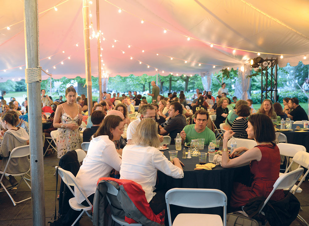 Guests seated at round tables in folding chairs under a white tent with white Christmas lights.