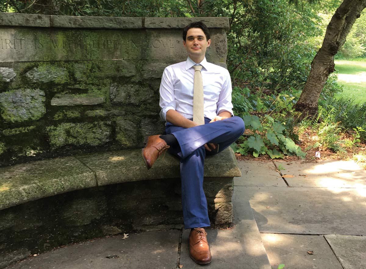 Joshua Sokol sits on a stone bench with one ankle crossed over his knee. He wears a white shirt, cream-colored tie, blue pants, and brown shoes.