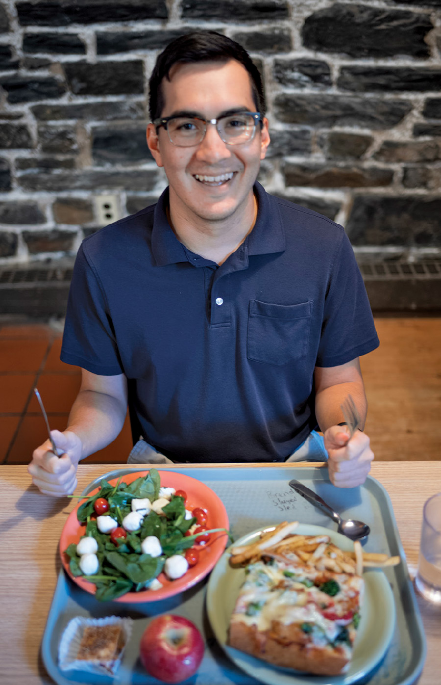 Alum Roy Greim '14 smiles and poses with utensils in hand, ready to enjoy salad, pizza, fruit, and fries.