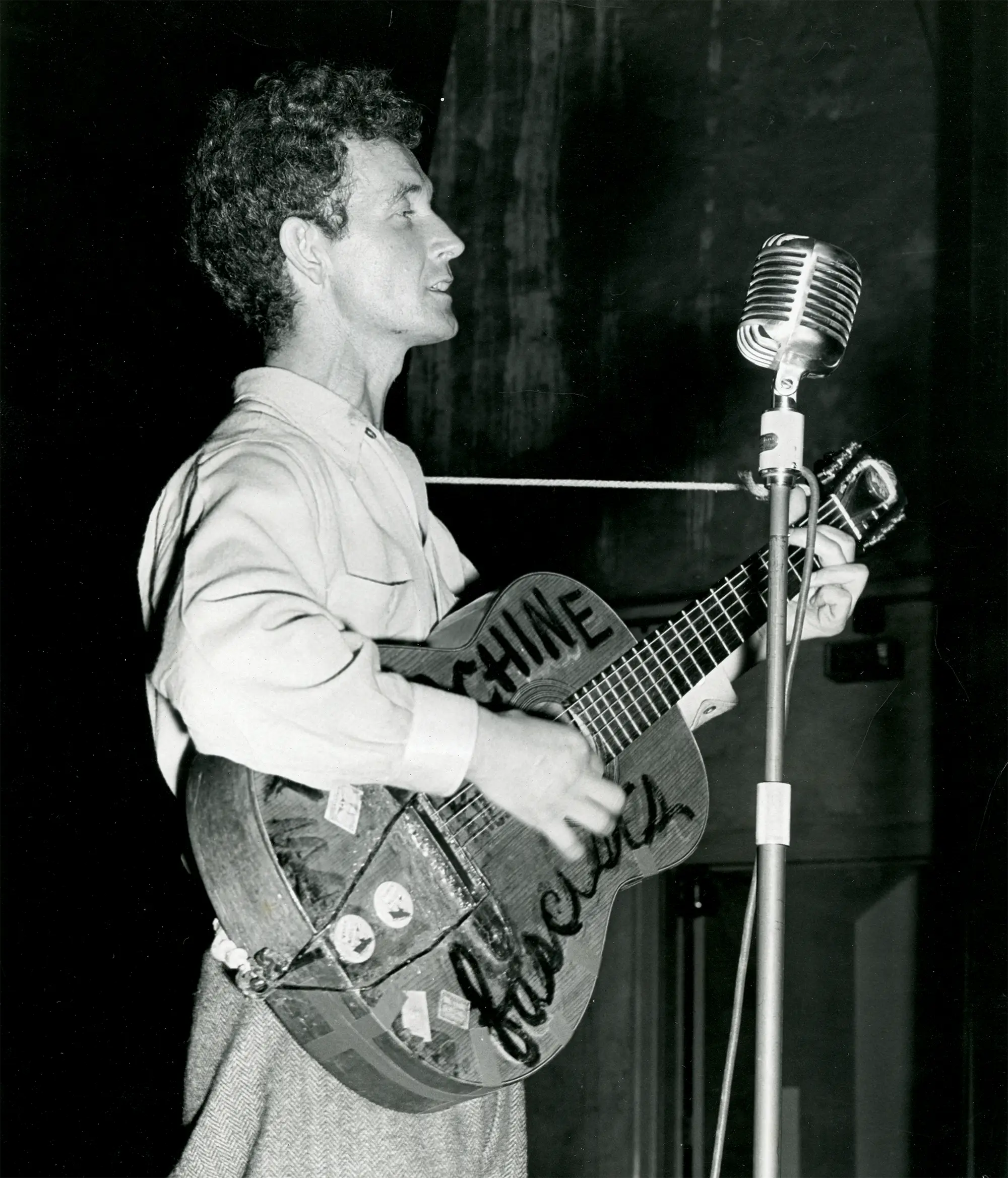 Black and white photo of Woodie Guthrie performing on his acoustic guitar with word This Machine Kills Facists in black lettering on the guitar.