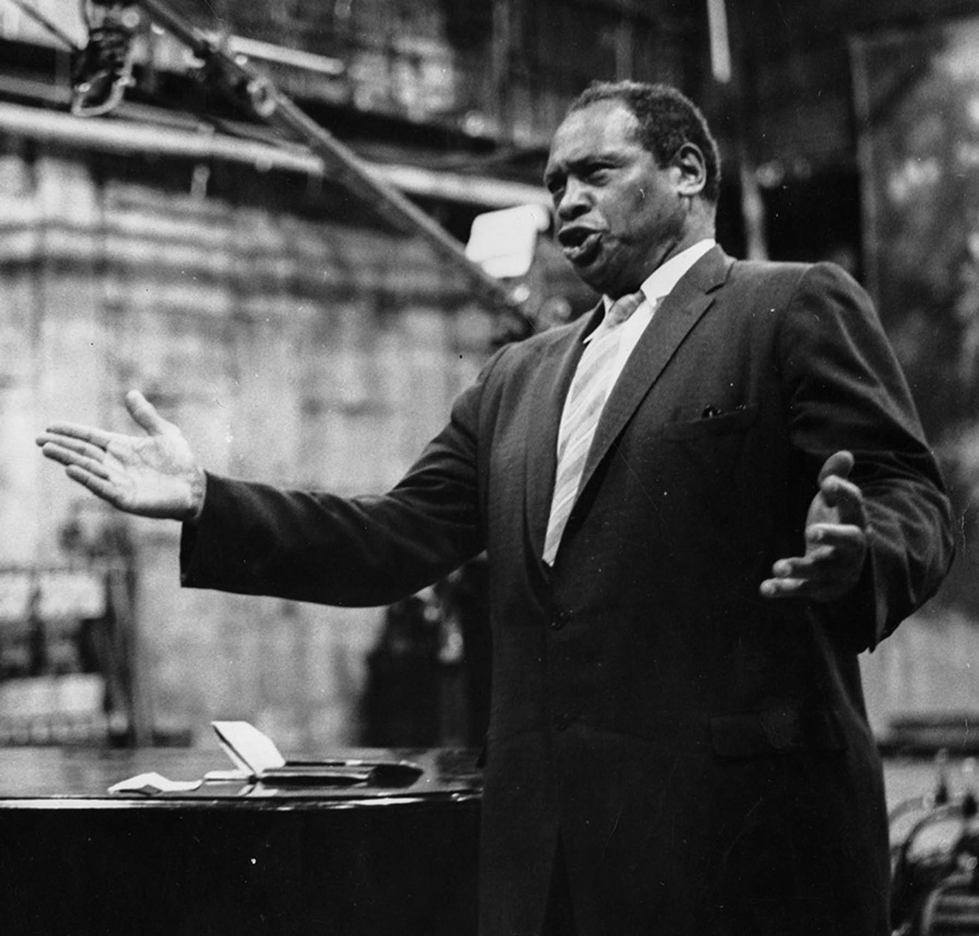 Medium long shot of Paul Robeson belting out tunes, standing, in black and white.
